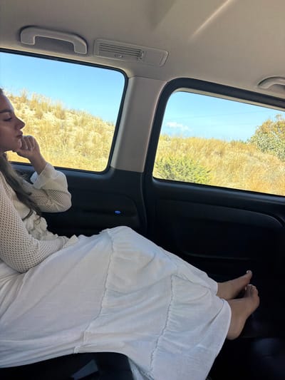 A person, presumably a female, is seen lying horizontally within a vehicle. The individual is wearing white clothing. A glimpse of the landscape outside the car window shows arid vegetation under a clear, blue sky. The interior of the vehicle reveals black panels, air conditioning vents, and a grab handle. The exact location in Thira Municipal Unit, Greece, is not discernible from the visual information.