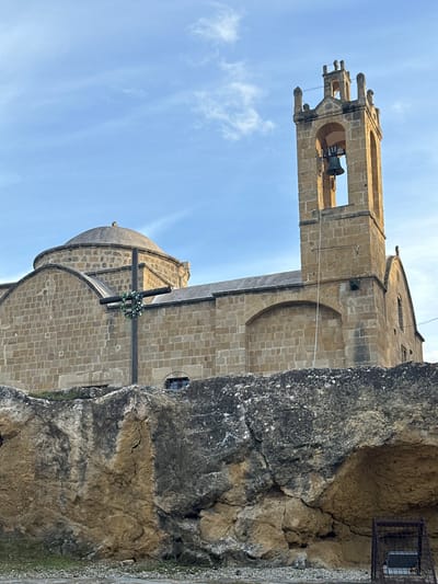 The image displays a stone church or monastery with a prominent bell tower and multiple domes, set against a bright, clear blue sky with wispy white clouds. The building is constructed from light-colored, rectangular stone blocks.

The bell tower, located on the left, is tall and features arched openings near its summit, through which a large bell is visible. The top of the tower includes decorative stonework. Further along the building, multiple domes with arched bases are apparent. In front of the church structure, a large wooden cross stands, adorned with a white floral wreath.

A rugged, natural rock face or cliff forms the right side of the scene, appearing to be adjacent to or partially integrating with the church's foundation. A small, dark metal cage or structure is attached to the upper right portion of this rock face.

The scene is illuminated by bright daylight, suggesting it is either morning or afternoon. No specific activity is taking place, and there are no visible people or discernible text within the frame. The overall impression is that of a serene and historical religious site, characteristic of the Mediterranean region, likely Agios Dometios, Cyprus, given the context.