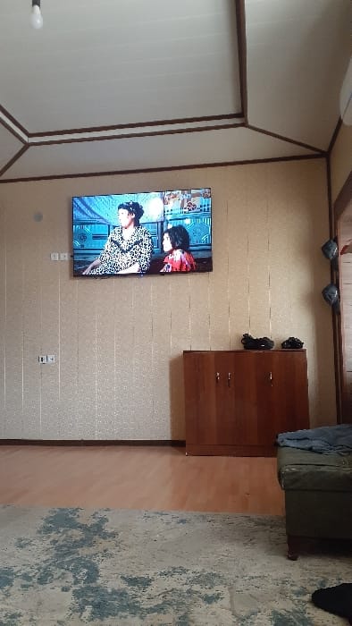 The image depicts an indoor scene within a room located in Pap, Uzbekistan. The room features light-colored, vertically paneled walls with a subtle sheen, a white paneled ceiling with dark brown trim, and a floor composed of light brown wood or laminate partially covered by a patterned area rug in cream and blue/grey tones. A large, wall-mounted flat-screen television is centered on the primary wall, displaying two female figures from a program: one in a leopard print top and another in a red patterned top. Below the television, a dark brown wooden cabinet with three doors and small metal handles is positioned against the wall. Two dark-colored objects, possibly electronic devices, rest on the cabinet's surface. Two white square electrical outlets/switches are visible on the left wall below the TV, with a single one further down. A bare incandescent light bulb hangs from the ceiling in the upper left. A dark green upholstered footstool or ottoman, draped with dark fabric, is partially visible in the lower right foreground. No individuals are present within the physical room.