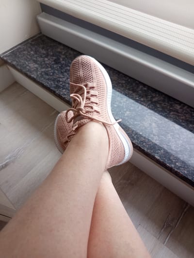A pair of light pink, knitted athletic shoes are worn by a person. The shoes have white soles and laces. The person's legs are bare and extend from the shoes. One leg is crossed over the other. The person is seated on a wooden floor with a granite-patterned ledge behind them. A light-colored blind is visible at the top of the frame. The scene appears to be indoors, possibly near a window. No time of day or weather is discernible. No text is visible.