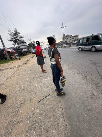 A public street scene displays two individuals. One is a taller woman wearing blue jeans, a gray top, black Crocs, and carrying a white purse. The other is a shorter person in a red shirt and gray skirt. They are standing on the edge of a paved area, near a road where vehicles are moving. Buildings and a cloudy sky are visible in the background, adding to the urban atmosphere.