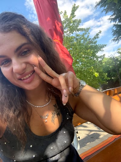 The image depicts a young woman posing for a selfie in an outdoor setting in Hrishteni, Bulgaria, under a bright blue sky with white clouds. The woman, with long brown wavy hair, is smiling broadly at the camera, revealing her teeth. She extends her left hand, making a peace sign with manicured fingernails featuring a light base and black outline design. She wears a silver-toned watch on her left wrist. Around her neck are two necklaces: one of white beads/pearls and another silver-toned chain with a floral cluster pendant and a clear teardrop charm. A floral tattoo is visible on her left upper chest. She is wearing a dark, ribbed tank top adorned with small, shiny studs or rhinestones. The background includes green foliage from trees, some bearing small red berries, and a large red fabric element, likely an umbrella or awning, providing shade. Wooden structures and benches are visible to the right, with sunlight casting shadows on a paved surface.