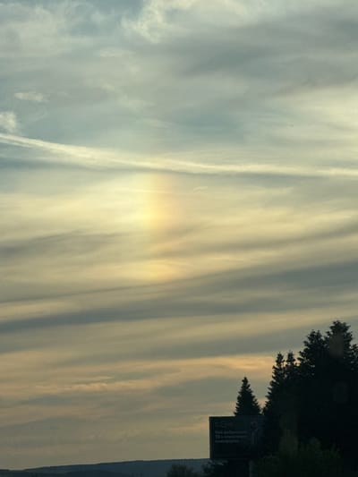 The image shows a natural phenomenon of a sun pillar, which appears as a vertical shaft of light extending upwards from the sun. The sun pillar is colored in pastel shades of yellow, orange, and pink, with a rainbow effect. It is visible in the sky above Gabarnitsa, Bulgaria, over a landscape of rolling hills and trees. The sky is filled with wispy clouds, and the overall atmosphere is serene and peaceful. There are no people or specific objects in the immediate vicinity of the sun pillar, but in the lower right corner of the image, there is a billboard with some text in Cyrillic script, and a silhouette of trees.