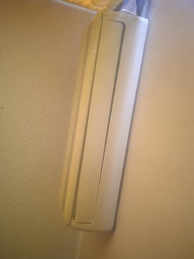 A light-colored air conditioner unit is mounted vertically on a wall. The wall on the left is textured and appears to be painted a pale yellow. The air conditioner unit is smooth and has a subtle ribbed design along its front. A dark grey conduit pipe is visible extending from the top of the unit, connecting it to something above. The wall on the right is a solid, lighter beige color. The lighting suggests an indoor environment, possibly a room or hallway. The scene is located in Puerto La Cruz, Venezuela.
