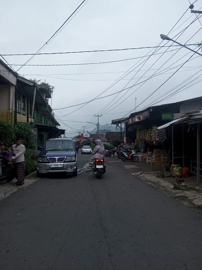 This image depicts an overcast daytime street scene in the city of Houaythat, Laos. On the left side of the asphalt road, a dark blue Mitsubishi SUV with license plate F 1556 YZ is parked next to a multi-story building featuring green trim and upper-level vegetation. A man in a light-colored shirt and plaid sarong stands near a black motorcycle, holding a small child, with another child partially visible beside them. In the center of the road, a person wearing a light purple head covering and patterned clothing rides a black motorcycle with license plate F 6872 WBN away from the viewer. Further down the street, a silver car is also moving away. The right side of the road is lined with single-story commercial structures, one displaying a sign reading "MINIGOLA AL-BUSTOMI," with merchandise hanging outside and cardboard boxes stacked on the ground. Several motorcycles are parked near these shops, and two large sacks, one green and one orange, are on the ground. Numerous utility poles with a dense network of overhead wires crisscross the frame. In the distant background, hills and an antenna tower are visible beneath the gray sky, with a street light fixture extending into the upper right corner.