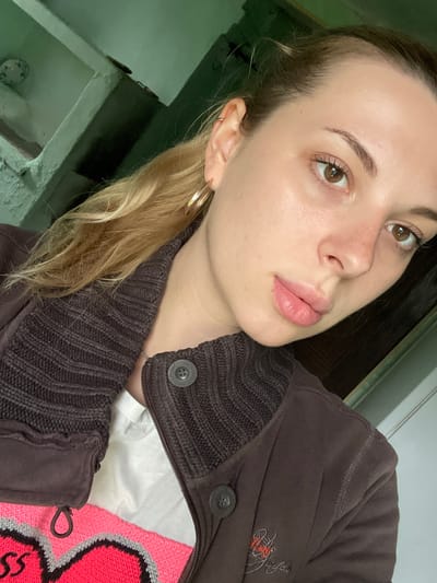 The image is a close-up selfie of a young Caucasian woman with blonde hair pulled back. She is wearing a brown zip-up jacket over a white t-shirt with a bright pink and grey heart design. She has light brown eyes and light skin with some freckles. The background appears to be an indoor setting with green painted walls and shelves, suggesting a modest living space. The overall impression is casual and informal.