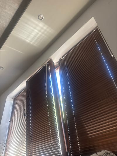 The image shows a room with blinds on a window. The blinds are brown and appear to be made of wood or a wood-like material. The blinds are partially open, allowing sunlight to enter the room and cast patterns on the ceiling. There is a recessed light fixture on the ceiling. The image is taken from a low angle, looking up towards the window and ceiling. There are no people or animals visible in the image. The location is specified as Belozem, Bulgaria.