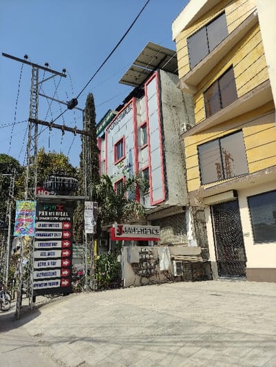 The image displays a bright daylight street scene in Rawalpindi, Pakistan, captured from a slightly elevated perspective looking towards several urban buildings. In the foreground, a paved surface leads to a cluster of multi-story structures. One building exhibits a distinctive yellow facade with dark, reflective windows, while an adjacent one features a light gray exterior highlighted with red trim around its windows. Solar panels are visible on the roof of one of the buildings. To the left, a prominent utility pole laden with numerous overhead wires stands against a clear, sunny blue sky.

A large, vertical sign is prominently displayed on the right side of the frame, advertising "PAN MEDICAL & DIAGNOSTIC CENTRE." Beneath the main title, a list of services is presented with red downward arrows: "AESTHETICS," "EMERGENCY 24/7," "LABORATORY," "ICU, MICU," "ECHO, K-RAY," "ULTRASOUND," and "OPERATION THEATER." Adjacent to this primary sign, another sign in red lettering reads "UZAESTHETICS." The overall scene depicts a static, commercial or mixed-use urban environment under a clear, sunny sky, with no human activity immediately visible.
