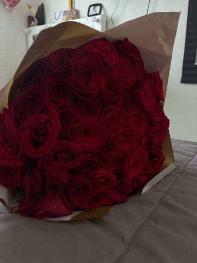 A large bouquet consisting of numerous red roses, tightly arranged and partially wrapped in brown and white paper, is centered in the foreground. The bouquet rests on a grey, quilted surface, likely a bed or sofa. In the background, against a light-colored wall, a white dresser or cabinet is visible. On this surface, indistinct objects including a dark-framed item, a white cable, and other small personal effects are present. A partially visible red heart-shaped balloon is in the upper left corner, and a dark window blind or shutter is on the far right. The scene is indoors in Komchén, Mexico. No individuals are present.