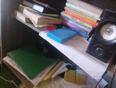 The image shows a cluttered bookshelf in a home or office located in Rawalpindi, Pakistan at the coordinates 33.498, 73.145.  A large black speaker is positioned in the lower right corner of the shelf, with a white paper label and a black grill in front. Several stacks of books are on the shelf, primarily written in Urdu. One book is open and laid flat on the shelf. A small, blue rectangular object with an inscription in Urdu sits on the shelf next to the books. A stack of paper and a notebook with a green cover lay on top of the books. The bottom of the image reveals a section of the floor, covered in green carpet and electrical cords.  Several papers, including a yellow sheet with a blue inscription, are lying scattered on the floor below the shelf.