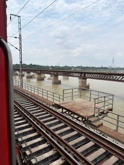 The image shows a view from a train window, looking out over railway tracks towards a large, steel truss bridge spanning a muddy river. The bridge has multiple spans supported by concrete piers. In the distance, there are trees and buildings, including power lines and towers. The sky is cloudy, with patches of blue. The railway tracks in the foreground are made of steel rails and concrete sleepers. A red train car is visible on the left side of the frame, partially obstructing the view. There is a metal platform with a railing and stairs next to the tracks, possibly a maintenance or inspection area. The location is identified as Shuklaganj, India.