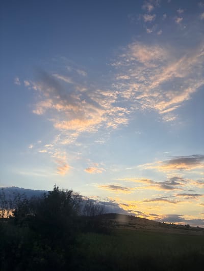 The image depicts a sunset over a rolling landscape. The sky is a gradient of blues, transitioning to soft oranges and pinks as it nears the horizon. Wispy clouds are scattered across the sky, catching the warm light of the setting sun. The landscape below is characterized by undulating hills covered in dry grass, with scattered trees and bushes. The foreground is dominated by darker vegetation, possibly fields or dense undergrowth. The scene is devoid of any human presence or activity. The location is identified as Blatnitsa, Bulgaria.