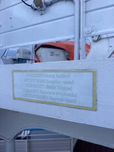 A sign with a yellow border is affixed to a white metal structure. The sign contains text in multiple languages, warning of a steep ladder. The text reads: "CAUTION! Steep ladder!", "ATTENTION! Escalier raide!", "VORSICHT! Steile Treppe!", "CUIDADO! Escalera empinada!", and "ОСТОРОЖНО! Крутой трап!". Behind the sign, an orange object with white text reading "30" is visible, possibly a life raft. The white metal structure appears to be part of a ship or boat. The background is blurred, but suggests an outdoor, possibly maritime environment. There is no specific indication of the location being in the city of Imeni Sverdlova, Russia.