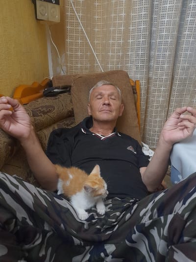 A man is lying down on a brown sofa, possibly in a living room. He appears to be sleeping or resting with his eyes closed and a faint smile on his face. He is wearing a black polo shirt with a red and white striped collar and what looks like an Adidas logo. A small, orange and white kitten is curled up on his chest, also appearing to be asleep or resting. The man is covered by a camouflage-patterned blanket or clothing.
In the background, there are light-colored curtains with a patterned texture. To the left of the man, on a yellow surface, sits a black object with two circular indentations, which might be a small speaker or some electronic device. A white cord is connected to this object and runs towards the right side of the frame.
The overall scene suggests a moment of quiet relaxation and companionship between the man and the kitten. The lighting appears to be indoor, possibly from a window, indicating it could be daytime. No specific weather cues are visible. The setting seems to be a home environment. There is visible text on the man's shirt which appears to be an Adidas logo.