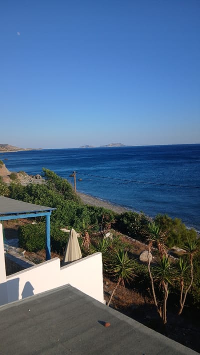 This media file shows a view of the Aegean Sea from a slightly elevated perspective, likely a balcony or rooftop, in Kerames, Greece. The image is oriented vertically. In the foreground, there is a dark grey rooftop and a portion of a white structure, possibly part of a building. Beyond the structures, there is lush green vegetation and several yucca plants with spiky leaves. A narrow, pebbled beach borders the vibrant, deep blue sea. The water stretches out to the horizon, where a distant, hazy coastline is visible. The sky is a clear, bright blue, with a faint crescent moon visible in the upper left quadrant of the image. The lighting suggests it is daytime, likely late morning or early afternoon, with the sun casting distinct shadows. There are no people or visible text in the image. The overall scene conveys a sense of tranquility and natural beauty.