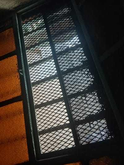 The scene depicts an interior view looking outwards through a secured window located in Ongata Rongai ward, Kenya. The foreground is dark, framing a vertical window structure composed of dark-colored metal. To the left, a section of orange-amber, textured glass with an irregular, possibly pebbled or hammered pattern, occupies approximately one-third of the window's width. The central and right portions of the window are fitted with two panels of dark, diamond-patterned expanded metal mesh, serving as security grates. The upper mesh panel exhibits multiple rectangular cutouts or sections with a denser or different mesh pattern. Bright light from an exterior source penetrates through the security grates, illuminating the diamond pattern and creating significant contrast with the darker interior elements. No individuals are visible. No specific actions or interactions are depicted. The environment visible through the grates is indistinct due to the mesh and lighting, showing only diffused lighter areas suggesting an exterior space.