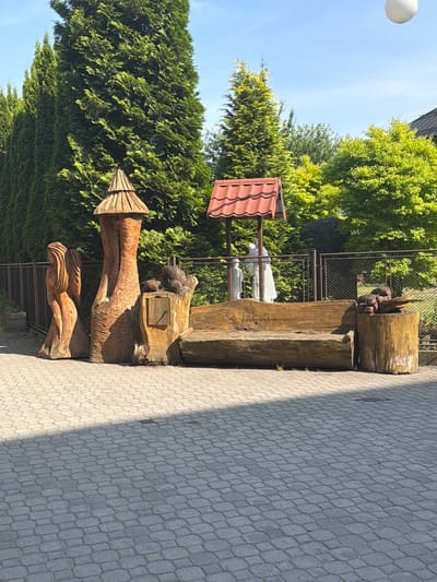 In Jablunkov, Czechia, a paved courtyard features a rustic wooden bench carved into a log. The bench is adorned with carved text reading "JACKI". To the left of the bench, a carved wooden statue of a figure with flowing hair stands next to another carved wooden structure resembling a small tower or stump with a conical roof. To the right of the bench, a cylindrical log serves as a side table, topped with what appears to be a carved animal or wood sculpture. In the background, a fence separates the courtyard from a row of tall, lush green evergreen trees. Behind the fence, a small, red-roofed structure stands, and two figures, possibly statues, are visible beneath it. Sunlight casts shadows across the paved area.