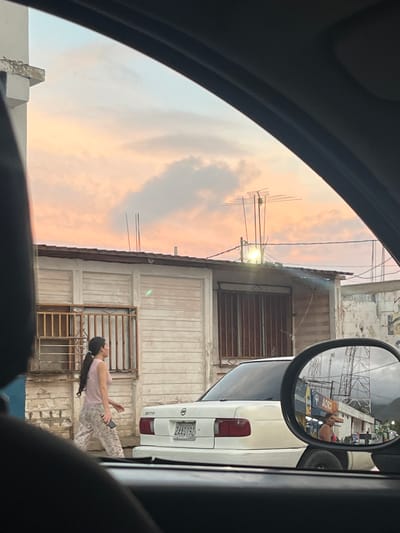 This image captures a street scene in Conuco Viejo, Venezuela, likely during the late afternoon or early evening, given the warm hues in the sky. The perspective is from inside a vehicle, looking through the windshield and side mirror.
In the foreground, a woman with long dark hair pulled back in a ponytail is walking from left to right, away from the camera. She is wearing a light pink tank top and patterned pajama-like pants. She is holding a dark object, possibly a phone, in her right hand.
To her right, a white sedan is parked. The car appears to be a Nissan Sentra, with a Venezuelan license plate reading "2AA0190".
The background features a low-rise building with light-colored wooden siding and barred windows. Above this building, a television antenna is visible against a sky with scattered clouds tinged with orange and pink. Further to the right, the side mirror reflects a street scene with buildings, possibly shops, and a person standing near one of them. In the far distance, hills or mountains are faintly visible. The overall environment appears to be a somewhat modest urban or semi-urban setting.