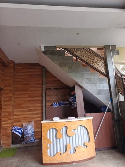 The image shows an interior space with a staircase and a reception desk. The staircase is constructed of gray stone, with a decorative railing featuring rock-like patterns. Next to the wooden wall, there's a front desk with a white and brown drip design. A plastic bag filled with what appears to be garbage sits on the floor next to the wall. A blue mop leans against the wall next to the stairs.