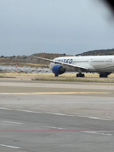 The image depicts a United Airlines 787-9 Dreamliner aircraft on a runway at Athens International Airport Eleftherios Venizelos, located in Spata-Loutsa Municipal Unit, Greece. The airplane is viewed from a side angle, revealing part of the fuselage, the left wing, and a blue engine. The runway is made of gray concrete with painted yellow and red lines. Beyond the runway is a dry field with rows of solar panels on a hillside. The sky is overcast and gray.