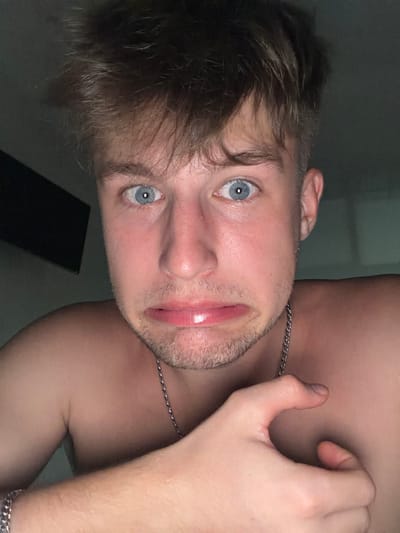 A close-up, head-and-shoulders portrait of a young man is shown. He has light brown, messy hair and striking blue eyes. His mouth is contorted into a humorous grimace, with his lips pursed. He is shirtless, wearing a silver chain necklace. His right hand is raised, with his fingers bent in an unusual position near his face. The background is dimly lit and appears to be an indoor setting. There are no discernible landmarks or environmental clues that would indicate a specific location within Vanino, Russia, or any city.