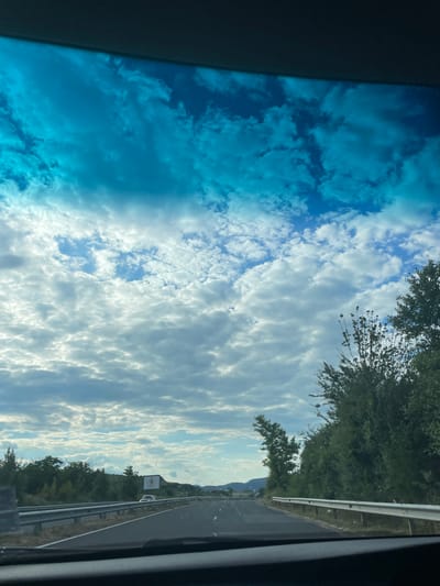 An outdoor, eye-level, full shot captured from a vehicle's windshield shows a highway with a bright blue sky and scattered white clouds. On the right side of the highway, there are green trees and vegetation. In the distance, there are hills and mountains, and a billboard with a circular image is visible. A white car is on the highway. The road appears to be asphalt and has a white line marking the lane.