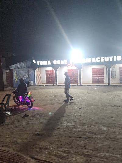 At night in Abraka, Nigeria, a man walks on the sidewalk in front of a building with the sign "YOMA CARE PHARMACEUTICAL" illuminated. A motorcycle with colorful lights sits near the curb in front of a building with corrugated metal sheeting. The ground appears to be a dirt or unpaved surface. Another person is next to the motorcycle.