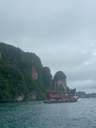 This is a photograph taken from a body of water, likely the sea, looking towards a coastline with dramatic limestone cliffs covered in lush green vegetation. The sky is overcast and gray, suggesting cloudy or potentially rainy weather. In the foreground, the water is a deep turquoise with visible ripples.
Dominating the mid-ground is a large, multi-decked wooden boat designed to resemble a pirate ship. The boat is dark brown and has several passengers visible on its decks, some standing and others appearing to be seated. The boat has multiple masts and rigging, and life preservers are visible along its railings. The name "P PIRATE" is visible on the side of the boat in white lettering.
To the left of the pirate ship, a smaller, dark-colored boat with at least one person visible is moving through the water. Further in the distance, more boats and landforms are visible. The limestone cliffs rise steeply from the water, with sheer rock faces interspersed with dense green foliage. The overall impression is a scenic, coastal landscape with a tourist boat. The location is Haad Hin Khom, Thailand.