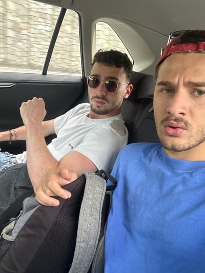 The image depicts two adult males inside the rear passenger compartment of a moving vehicle, identified as being in the city of Olgiate Olona, Italy.
The male in the foreground, on the right side of the frame, is partially visible from the chest up, suggesting he is operating the camera for a selfie. He has light skin, a red baseball cap worn backward, and is wearing a blue short-sleeved t-shirt. His lips are slightly pursed, and his eyes are directed towards the camera. His right hand is visible, resting on a gray and black backpack positioned between the two subjects.
The male seated in the rear left passenger seat is visible from the waist up. He has light skin, short dark curly hair, and stubble. He is wearing round sunglasses with gold-colored frames and dark lenses, a plain white short-sleeved t-shirt with a small dark symbol on the left chest, and dark jeans. A beaded bracelet is visible on his left wrist. His left arm is bent, with a clenched fist resting on his lap or side. His gaze is directed forward towards the camera.
The vehicle's interior is dark, with visible seating surfaces, a headrest featuring a geometric pattern, and a window frame. Through the left rear window, a blurred, light-colored external environment with horizontal lines is visible, indicating the vehicle's motion.