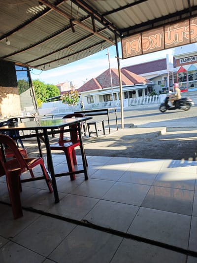 An outdoor seating area of a shop or restaurant is shown from a low angle, looking towards a street. In the foreground, a metal table with a reflective top is surrounded by red plastic chairs. The floor is tiled with light-colored squares. A corrugated metal roof, supported by rusty metal beams, extends over the seating area. Beyond the seating area, a street is visible with a white fence and residential buildings. A person on a scooter with a pink helmet is riding on the street. A sign with orange lettering is visible above the street, appearing to read "KOPI UTU" or similar. The overall scene suggests a warm, sunny day in a commercial area of Geudonggeudong, Indonesia.