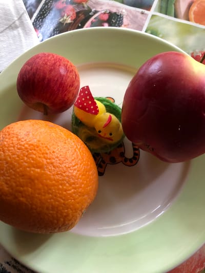 The image displays a plate with three pieces of fruit and a small, fuzzy toy chick. Two apples, one predominantly red and the other a mix of red and yellow, are positioned at the top of the plate. An orange rests at the bottom left. The toy chick, yellow with a red polka dot hat and a small red beak, is situated between the apples and behind the orange, nestled in green material that resembles straw or grass. The plate itself has a pale green rim. The background shows a surface with a patterned design, featuring images of berries and possibly other fruits, suggesting it might be a tablecloth or placemat. The scene is indoors, with no specific environmental features that can be used to determine a location in Staroe Istomino, Russia.