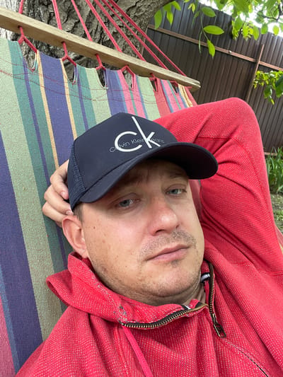A man in a red hooded sweatshirt and a black baseball cap with the Calvin Klein logo is lying in a striped hammock. The hammock is suspended from a tree. A brown wooden fence and green foliage are visible in the background. The scene appears to be outdoors, possibly in a backyard in Krutets, Russia.