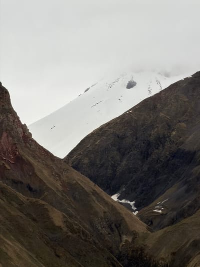 The image shows a low-angle view of a mountainous landscape. Prominent rock formations dominate the right side, with visible striations and textures. The left side displays a snow-covered or cloudy area. Small patches of snow are also visible on the rock faces.
