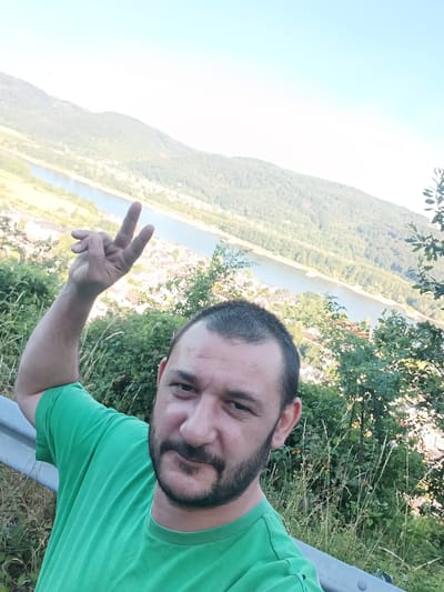 A man with a beard, wearing a green t-shirt, takes a selfie outdoors. He is holding up his right hand in a peace sign. The background shows a river valley with a town nestled on its banks, and rolling, tree-covered hills beyond. This scene is located in the vicinity of Leutesdorf, Germany. A metal railing is visible to the left of the man.