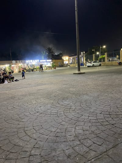 A night exterior shot shows a paved area with a circular pattern in front of a restaurant in Tipaza, Algeria. The restaurant has a lit sign that says "RESTAURANT" and "CHIKU GAMIS." People are sitting and standing in front of the restaurant and on the paved area. A child in a blue shirt and shorts is walking across the paved area. A white car is parked in the background. Streetlights are visible, illuminating the scene. The sky is dark.