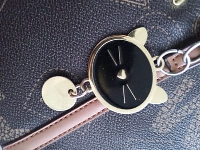 Location: Kabal, Pakistan.
This image presents a forensic, close-up view of a textured dark brown surface, resembling grained leather or a patterned canvas material, likely part of an accessory such as a handbag or wallet. Attached to this surface, via a light brown leather strap with visible stitching, is a metallic chain assembly featuring two distinct charms. The primary charm is a gold-toned circular piece with small, rounded ear-like protrusions at the top, enclosing a black enameled face. This black face displays two thin, vertical, light-colored lines on either side, and a smaller, pointed element at the center, stylized as a cat's face. This charm is connected by a gold jump ring to a series of interlocking silver-toned rings. A secondary, smaller, plain gold-toned circular disc charm is also attached to one of these silver rings. The dark brown background material exhibits a consistent pebbled texture interspersed with lighter, faded patterns. No individuals or actions are depicted; the focus is solely on the objects and their material details.