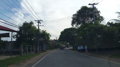 The image depicts a street scene in Meunasah Lhok, Indonesia. A paved road curves into the distance, flanked by grassy verges and dense foliage. Several utility poles with numerous overhead wires are prominent features in the landscape. On the left side of the road, a building with a red roof is visible, adjacent to a fence and more greenery. Several vehicles, including a dark-colored SUV, are present on the road. In the middle ground, three individuals wearing headscarves are walking along the right side of the road. The sky is overcast with scattered clouds.