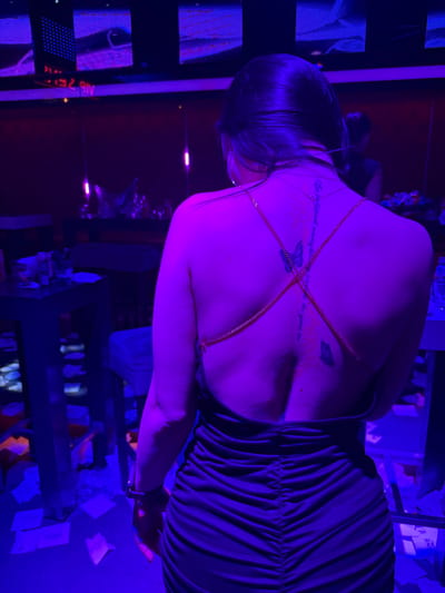 A woman is observed from behind, standing in a dimly lit indoor environment characterized by purple and blue illumination. She wears a dark, backless dress with ruching, and a gold-colored body chain is draped across her upper back. Visible on her back are tattoos: text oriented vertically along the spine and two butterfly motifs. A large hoop earring is present on her right ear, and a watch or bracelet is on her left wrist. The background features multiple rectangular screens displaying purple and blue light patterns. Dark, elevated surfaces resembling tables or bars are visible, holding indistinct objects. The floor is strewn with white papers. A blurred figure or reflection is distantly visible behind the woman. The specific location within Vaglarovo, Bulgaria, is not discernible from the image.