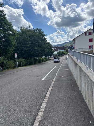 The image shows a street in Lausen, Switzerland, viewed from an elevated perspective. The street is asphalt with white painted parking spaces and a border of cobblestones along one side. A white minivan is driving away from the viewer on the street. A building with red shutters runs along the left of the road, with a concrete retaining wall and metal railing separating it from the street. A large tree borders the other side of the street. A road sign labeled "Privat" is visible along the street. In the background are distant buildings and a mountain range. The sky is blue with large cumulus clouds.