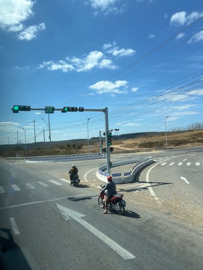 The image shows a road with two motorcycles stopped near a traffic light. The traffic light is green and displays the number 82. One motorcycle is red and the other is brown. There are directional arrows painted on the road. The sky is blue with scattered clouds. Power lines are visible above, and a grassy area is present in the background.