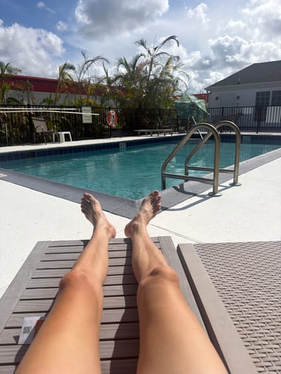 The image shows a first-person perspective of a person's legs and feet resting on a lounge chair next to a swimming pool. The pool is filled with blue water and has a metal ladder with handrails. A fence surrounds the pool area, with palm trees and a building visible in the background. The sky is partly cloudy. The scene appears to be outdoors, possibly at a hotel or residential complex in Palm Bay, Florida, United States of America.