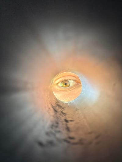 The image shows a close-up view of a human eye peering through the end of a cylindrical object, possibly a pipe or tunnel. The eye is the central focus, appearing large and detailed with a light brown iris and a dark pupil. The skin surrounding the eye is visible, with fine lines and pores. The cylindrical opening creates a framing effect, with the interior of the tube appearing textured and illuminated from within by a warm, orange light that casts shadows and highlights on the inner surface. The exterior of the tube and the surrounding area are dark and out of focus, creating a sense of depth and mystery. The overall mood is intense and intimate, suggesting a moment of observation or surveillance. There is no visible text in the image. The location context of Bašť, Czechia, does not provide further specific visual cues within the image itself.