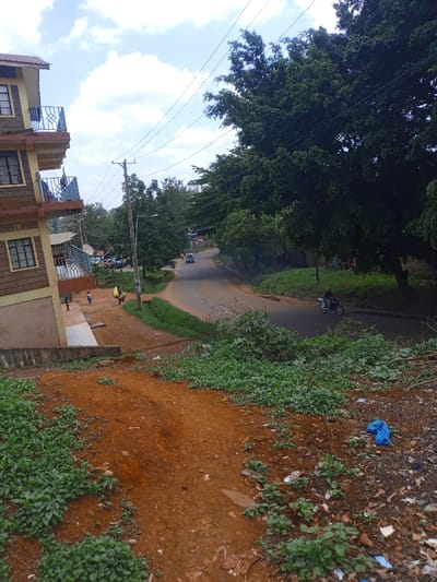 The image depicts a street scene in Murang'a, Kenya, viewed from an elevated position. The foreground consists of reddish-brown soil mixed with green ground cover and scattered refuse, including a blue plastic bag on the lower right.

To the left, a multi-story building with a light-colored facade and dark trim features several windows and blue-railed balconies. At the base of this building, a concrete retaining wall separates it from a dirt path. Several individuals, including children, are present on this path; one person is seen carrying a large yellow sack on their back, moving away from the viewer.

A paved, two-lane road curves through the midground, sloping gently downwards. It has unpaved dirt and grass shoulders. A wooden utility pole with multiple overhead power lines stands prominently near the road's edge. Adjacent to it, a metal pole supports a street light.

On the road, a dark-colored motorcycle with a single rider travels rightward. Further down the road, a light-colored sedan is visible. The right side of the road is lined with dense, mature green trees that extend into the background. Additional trees and bushes populate the left side of the road and the central area. The sky above is partly cloudy with white clouds against a pale blue background.