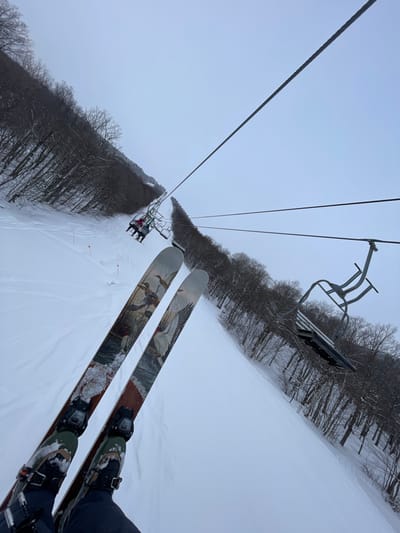 This first-person point-of-view image captures a scene from a ski lift ascending a snow-covered mountain at a ski resort, located in Jay, United States. In the foreground, the photographer's skis are prominently displayed, attached to ski boots. The skis feature distinctive artwork depicting ducks or other waterfowl.
Below the skis, a wide expanse of fresh, white snow covers the ski trail, with gentle tracks visible from previous skiers. The ski lift cables stretch upwards and into the distance against an overcast, light gray sky. An empty ski lift chair hangs directly above and ahead of the photographer. Further down the lift line, another chair is visible, carrying at least two individuals.
The ski slope is flanked on both sides by dense forests of bare, deciduous trees, indicative of a winter setting. The ground is uniformly covered in snow, suggesting cold, possibly snowy weather. The overall ambiance is that of a calm, cloudy day on the slopes. The primary activity depicted is riding a ski lift, preparing for or returning from skiing. No legible text is visible within the image.