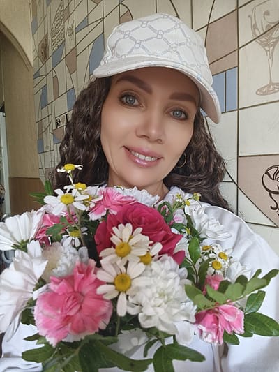 A medium shot captures a woman wearing a white baseball cap and holding a bouquet of flowers. She has long, dark, curly hair and blue eyes. She is smiling and looking at the camera. The background is a wall with a geometric tile pattern. The scene appears to be indoors, possibly in a residential setting or a cafe. The woman's clothing consists of a white shirt or jacket. The bouquet contains a mix of pink and white flowers, including roses and daisies. A ring is visible on her left hand. The overall lighting is bright and natural. The image is a selfie, indicated by the angle and the focus on the woman. There are no specific elements in the image that indicate the location as Chaikovsky, Russia.