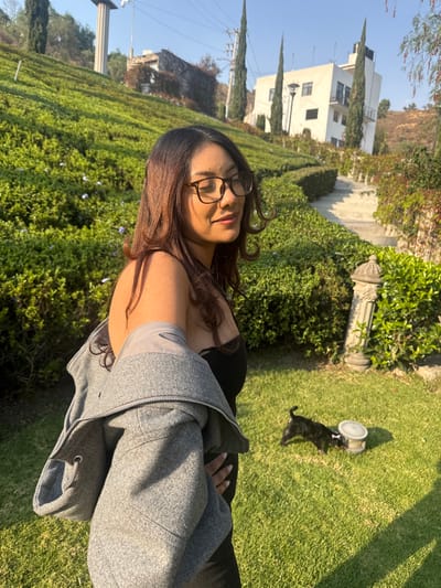 The scene is an outdoor, brightly lit garden area located within the city of Atizapán de Zaragoza, Mexico. A female, appearing to be in her twenties, with long, dark hair and wearing eyeglasses, stands in the foreground, facing slightly left with her eyes closed or squinting from direct sunlight. She is dressed in a black strapless garment, with a grey zippered jacket casually draped over her left shoulder. In the mid-ground, a small, dark-furred dog is positioned on a green lawn, attending to a light-colored bowl featuring a bone motif. The surrounding environment consists of lush, manicured green hedges and shrubbery, with a decorative stone pillar on the right. The background features a sloping hillside with a multi-story white building, tall slender trees, and a winding set of stone steps. A large, pale, columnar structure is visible on a more distant elevated area to the upper left, set against a clear blue sky.