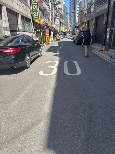 The image depicts a narrow urban street in Jongno-gu, South Korea, under clear, sunny conditions with distinct shadows cast across the asphalt. A large white "30" speed limit marking is painted centrally on the street in the foreground.
On the left side, a black Hyundai Grandeur HG 300 sedan is parked, facing away from the camera. Adjacent to it, building facades display various signs, including a prominent vertical yellow sign with red Korean characters reading "한의원" (traditional Korean medical clinic). Overhead electrical wiring and cables crisscross above the street.
On the right side of the street, a dark SUV is parked further down. A man, dressed in a dark short-sleeved shirt and light-colored trousers, stands facing away from the camera on the asphalt, holding a dark object in his right hand. A wavy yellow line is painted along the right curb. A red fire extinguisher box is visible on the sidewalk behind the parked SUV, and a light blue plastic chair is positioned further to the right. Additional vehicles are visible in the distance.