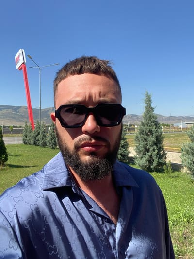 A man with dark hair, a beard, and sunglasses is taking a selfie outdoors in the city of Korkmaskala, Russia. He is wearing a blue patterned shirt. Behind him, there are trees, a red sign with white lettering on a pole, streetlights, a road, and hills in the distance. The sky is clear and blue, and the sun is shining.
