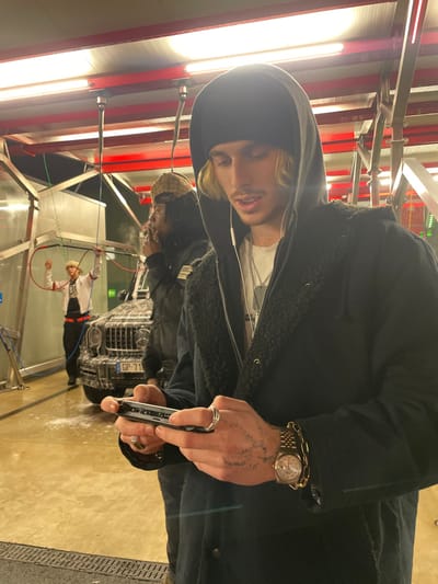 A car wash scene focuses on a fair-skinned individual in a black hooded jacket. Visible details include facial braces, headphones, a gold watch, and hand tattoos. The person holds a slim, dark object, possibly a phone or wallet. Behind them, a second person with patterned headwear stands near a car being washed. A third person appears to be actively cleaning the car. The background features bright lights and car wash equipment. The overall image seems to capture a casual, possibly social, moment at a self-service car wash.