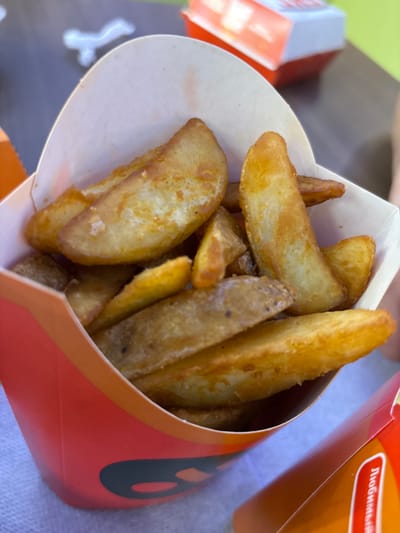 A close-up, eye-level shot shows a portion of potato wedges served in a red and white cardboard container. The wedges are golden brown and appear to be seasoned. The container has a printed logo with the text "Любимым" which is Russian for "Beloved". The background is blurred, suggesting an outdoor setting, possibly a park or street. The image is sharp and well-lit, focusing on the food.